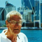 Elderly man wearing glasses at Sydney Harbour with skyscrapers in the background.